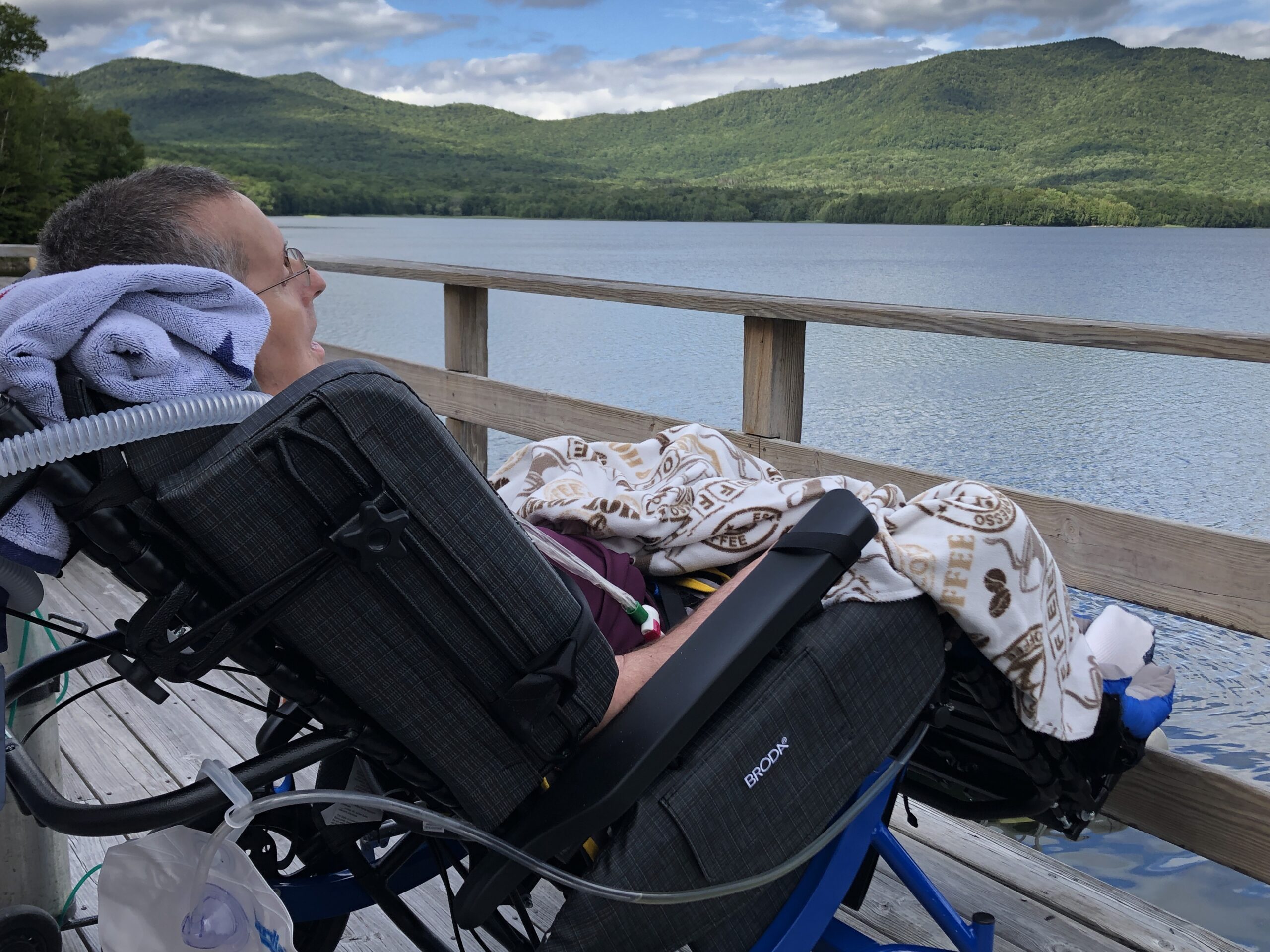 Man in burgundy shirt sits next to beautiful water scene with mountains in blue Synthesis Positioning Wheelchair