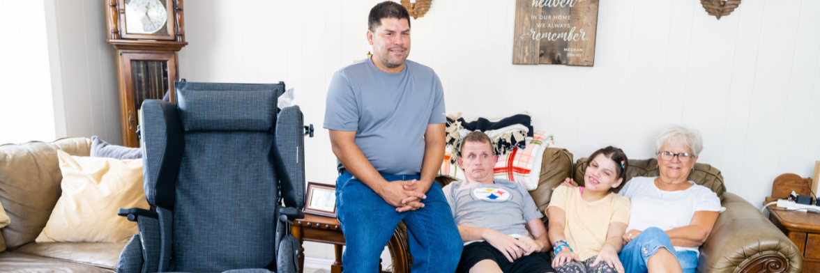 A family with Huntington's Disease sits on the sofa next to a Broda wheelchair.