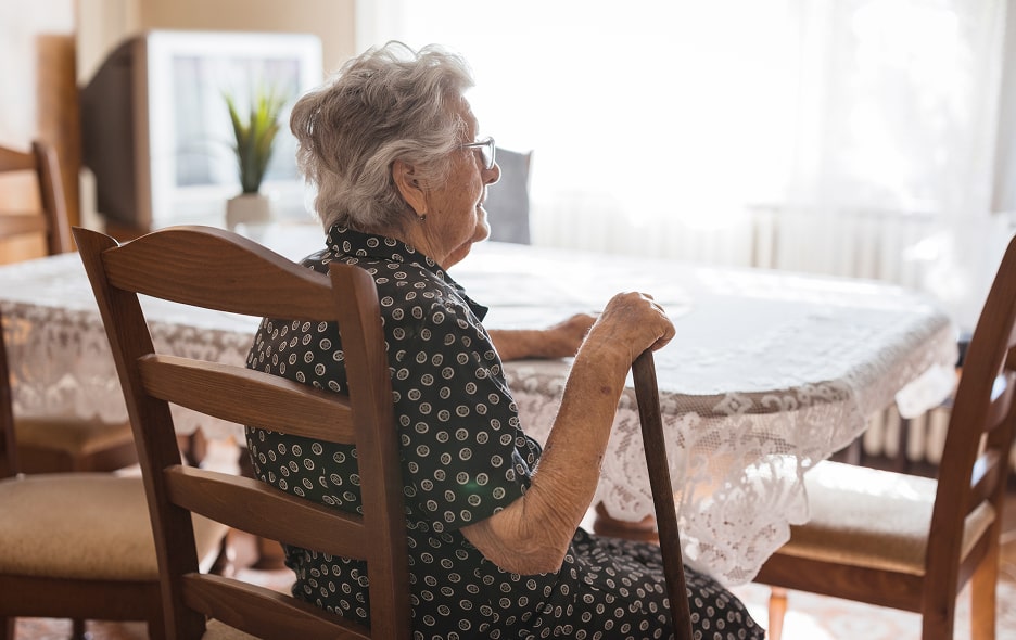 An elderly woman sits by a window with her cane