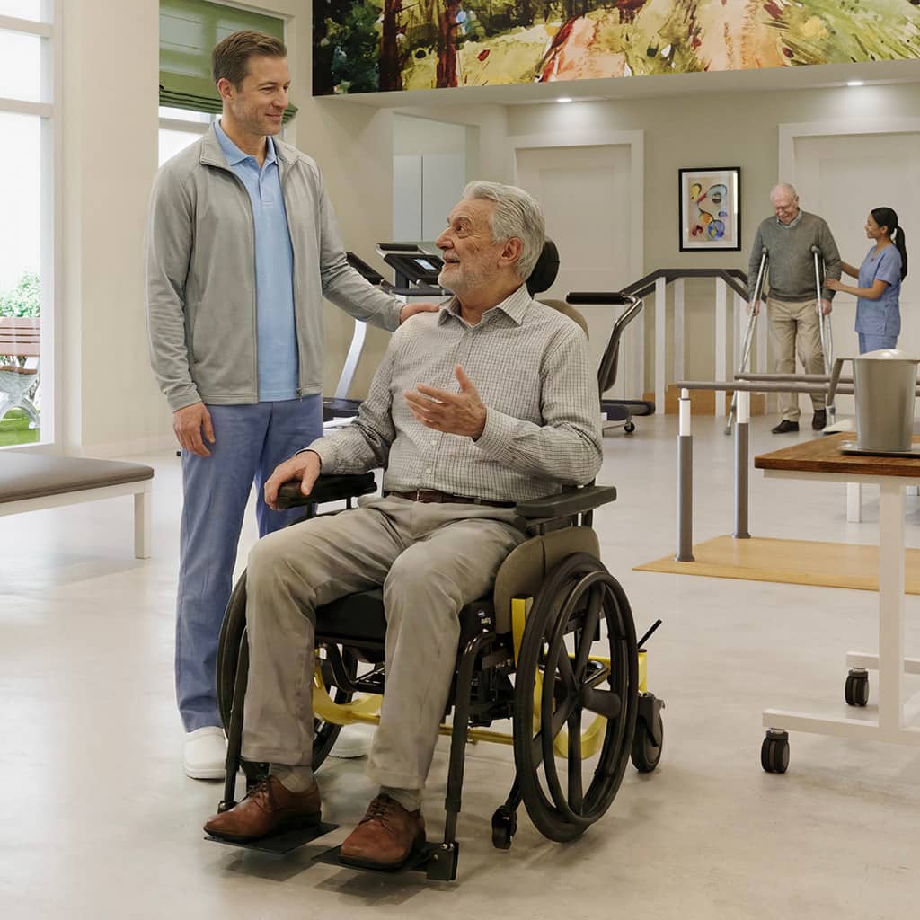 An older man in a wheelchair chatting with a young caregiver in the lobby